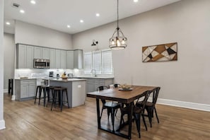 A modern kitchen with grey cabinets, a central island with stools, a dining table with chairs, and a hanging light fixture.