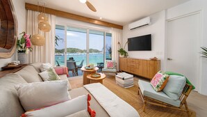Un salón amplio con un sofá grande, un ventilador de techo de madera, un televisor montado en la pared y vistas al mar a través de amplios ventanales.