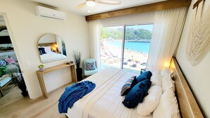 Un dormitorio con una cama grande, ventilador de techo, vista a una playa y una ventana con cortinas.