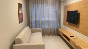 A modern living room with a beige sofa, a wooden TV stand, and a large window with sheer curtains. - Edifício Promenade II - Apto 403 (Maceio)