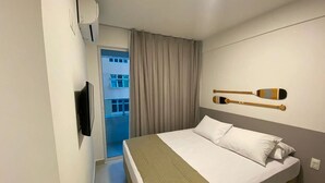 A hotel room with a bed, a television, a wall-mounted paddleboard, and a view of a building through a window. - Edifício Promenade II - Apto 403 (Maceio)