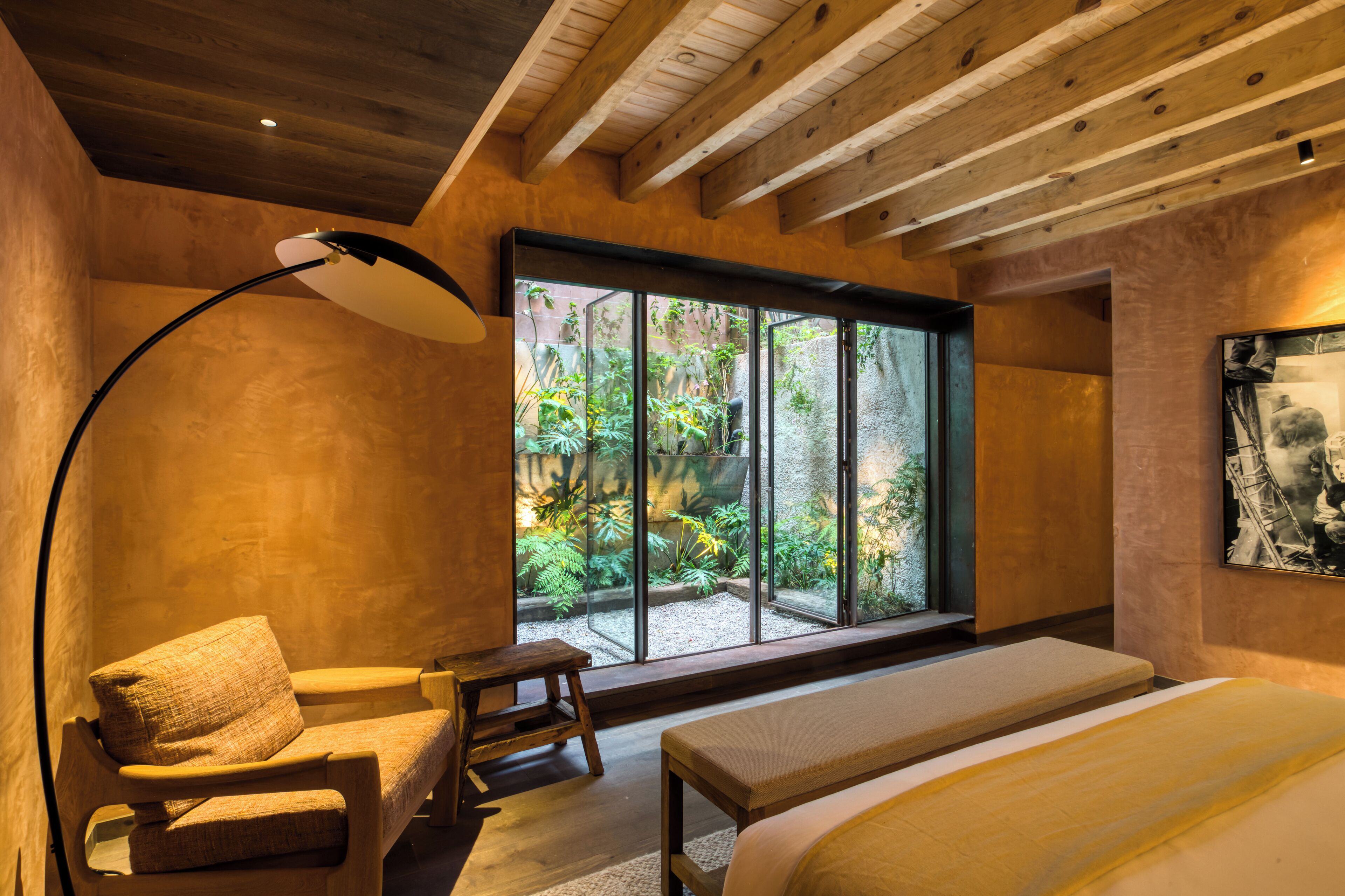 Quarto moderno com uma grande janela, vigas de madeira no teto, uma cama, uma cadeira e um abajur.
