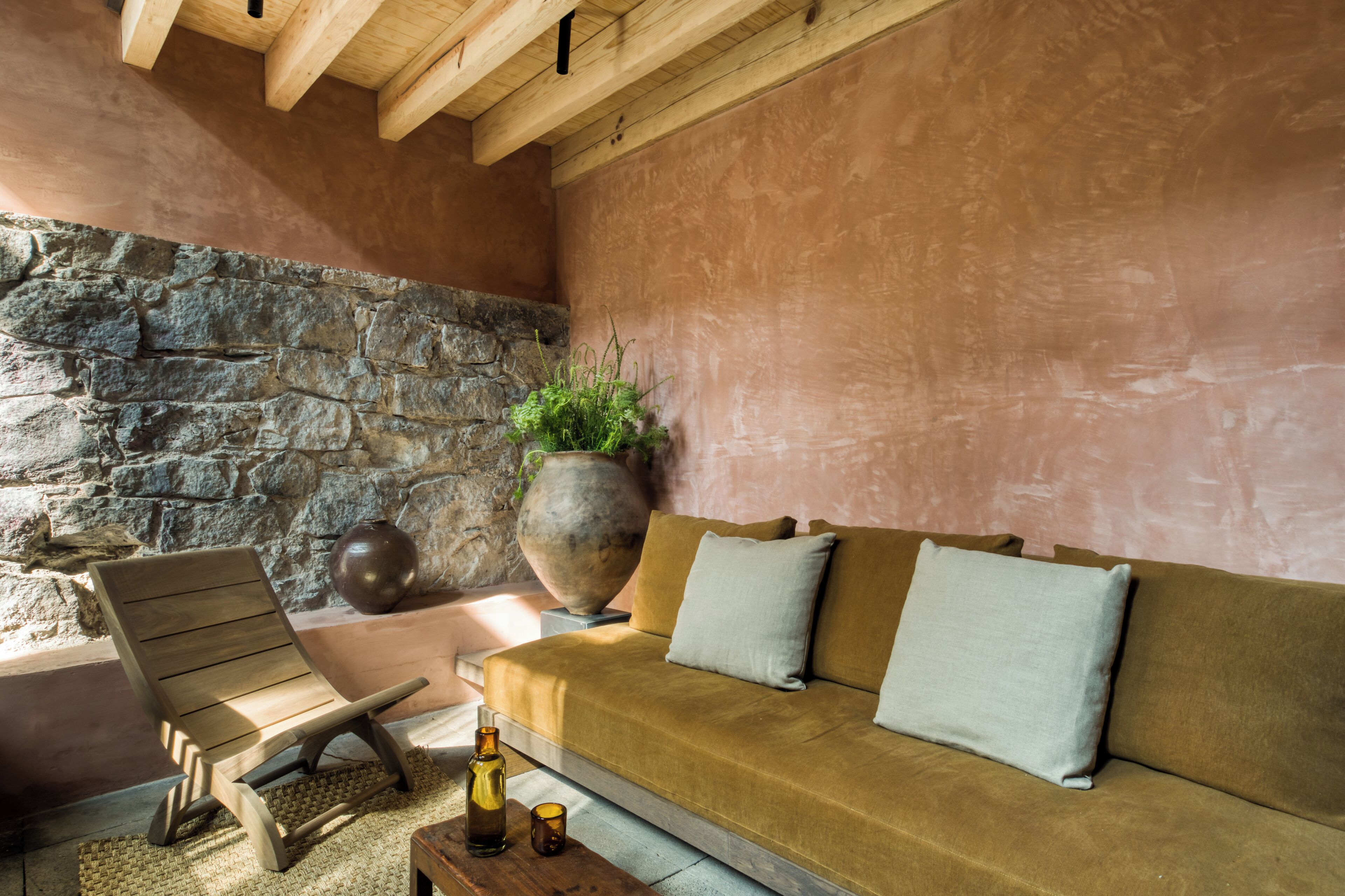 A cozy room with a stone wall, wooden ceiling beams, a sofa with cushions, a wooden chair, a large vase with a plant, and a small table with candles.