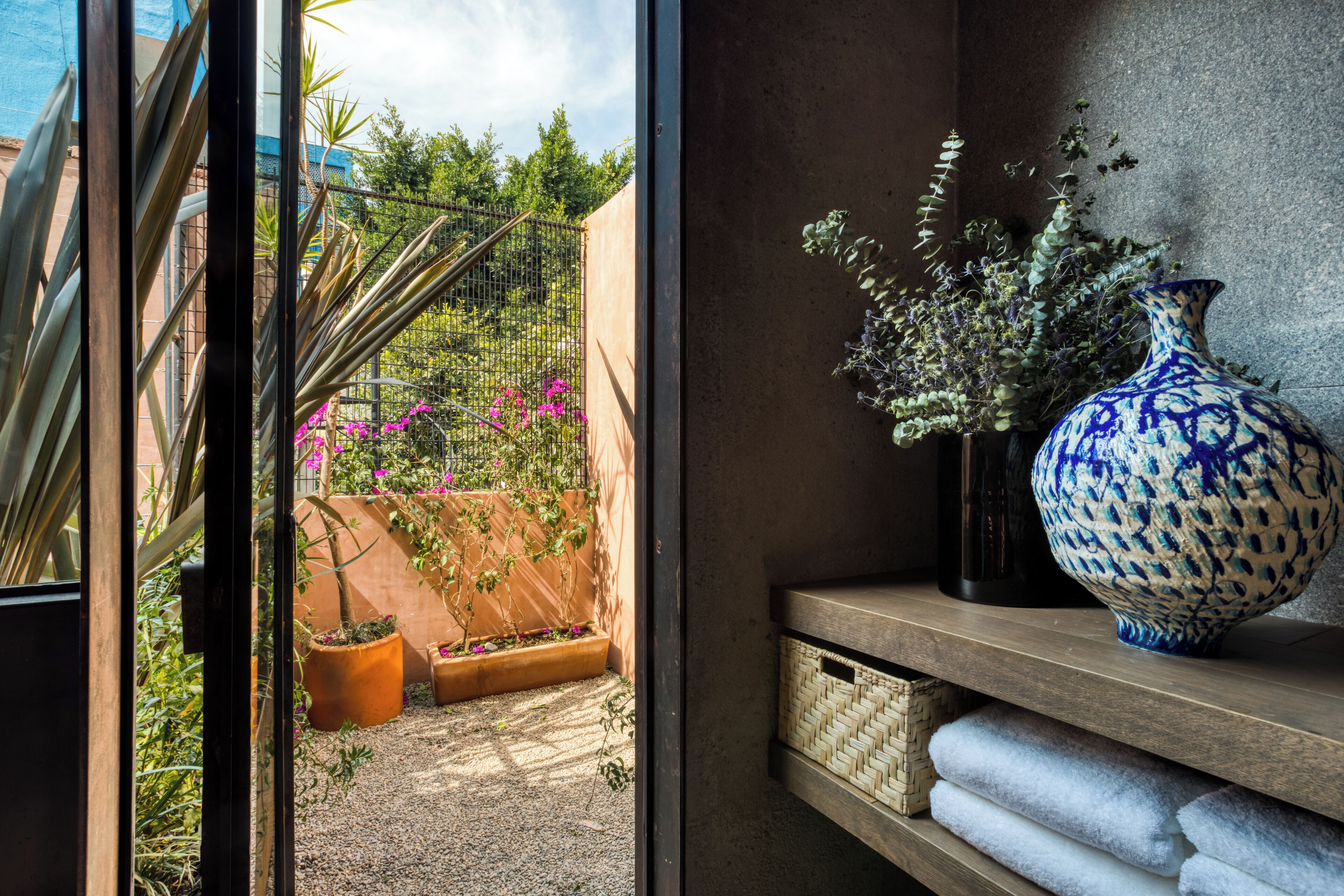 Quarto com uma grande janela com vista para um jardim, um vaso azul e branco numa estante, e uma cesta de vime.