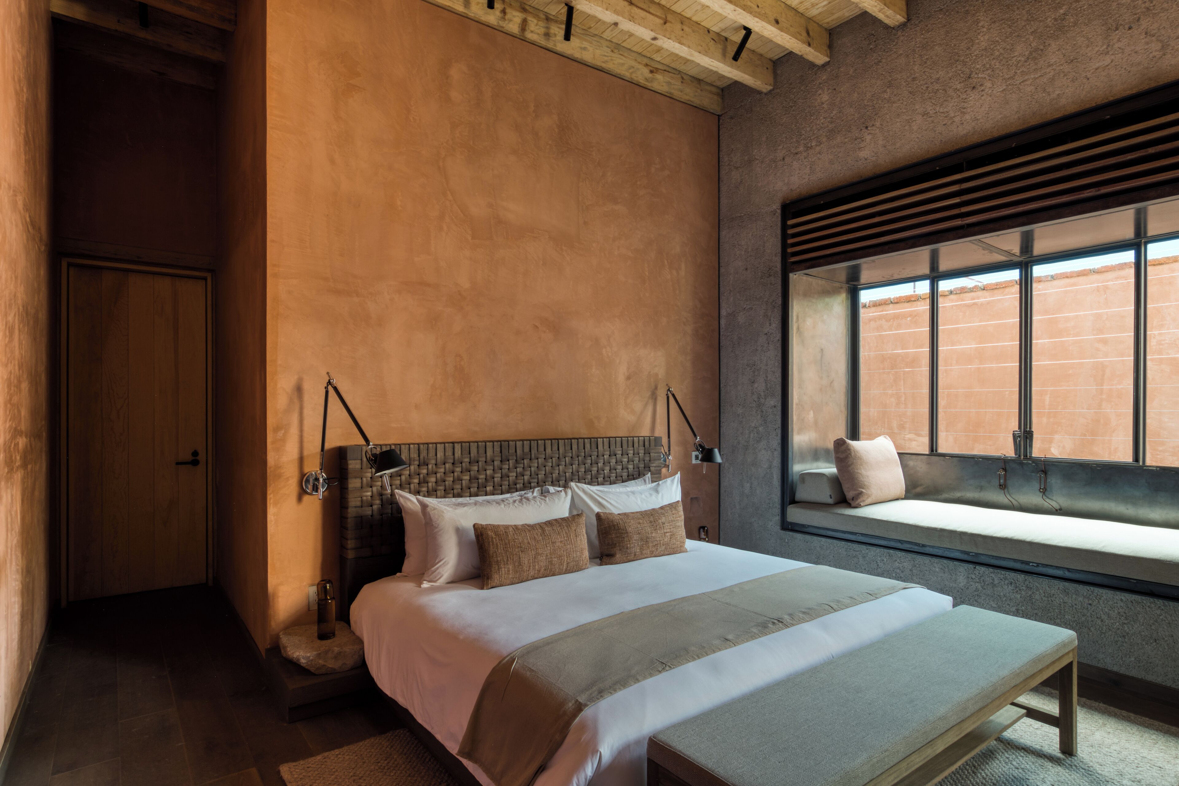 A bedroom with a large bed, a bench, a window with a view of a brick wall, and a wooden ceiling.