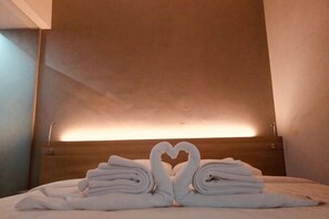A hotel room with a bed, a heart-shaped towel arrangement, and a wooden headboard.