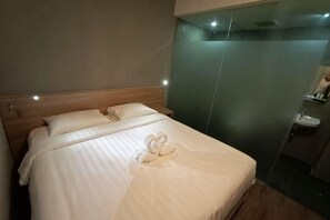 Habitación de hotel con cama, almohadas, toalla, cabecero de madera, pared verde y baño con lavamanos.