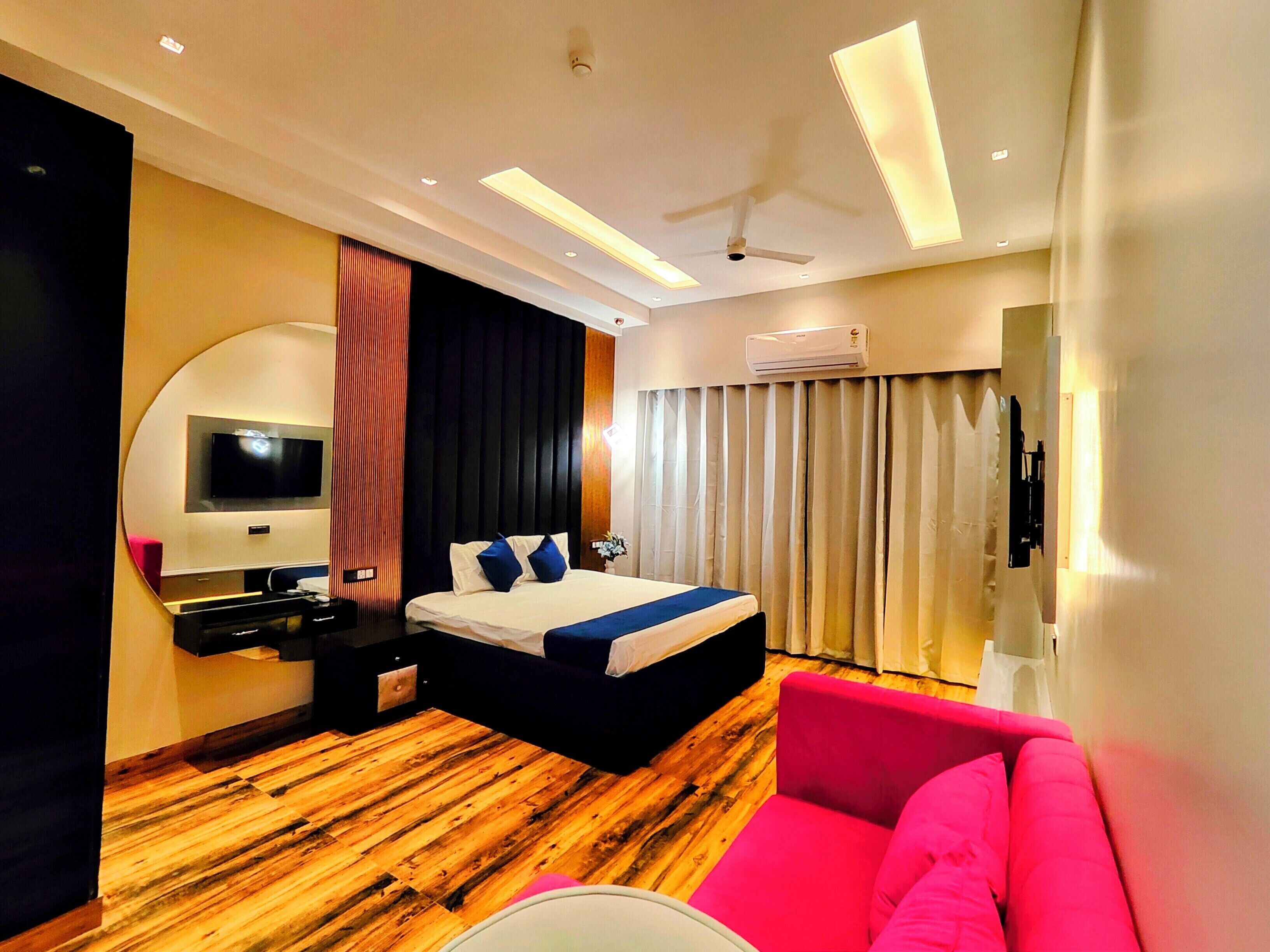A modern hotel room with a wooden floor, a bed with a blue accent, a pink sofa, a TV mounted on the wall, and an air conditioning unit.