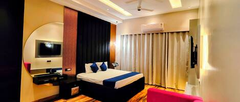 A modern hotel room with a wooden floor, a bed with a blue accent, a pink sofa, a TV mounted on the wall, and an air conditioning unit.
