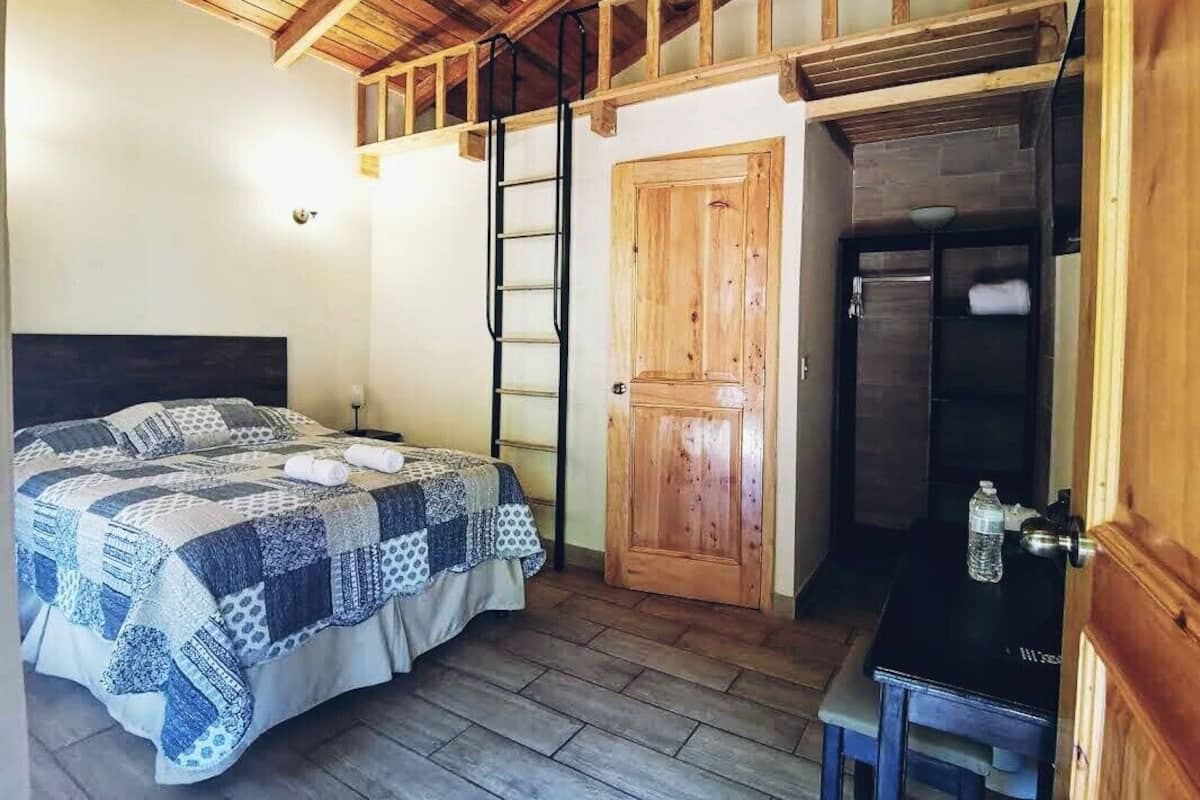 Un dormitorio con techo de madera, una cama con colcha, una puerta de madera y una mesita con una botella.