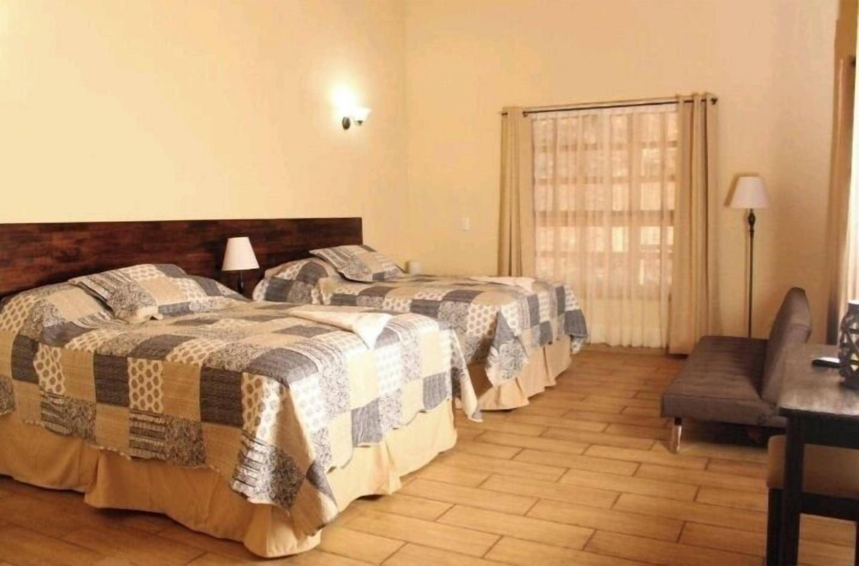 A hotel room with two beds, a wooden floor, a small table, and a window with curtains.