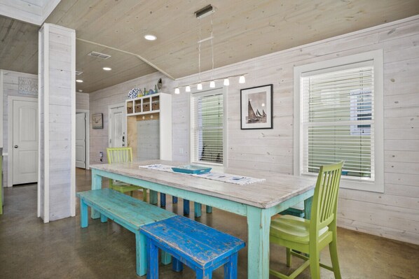 A dining area with a wooden table, benches, and chairs. There are windows with blinds, a ceiling with recessed lighting, and a wall with a framed picture.