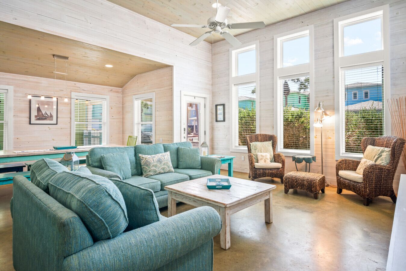 A spacious living room with a large sofa, a coffee table, and wicker chairs. There are large windows providing natural light and views of the outside.
