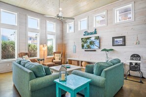 A spacious living room with a high ceiling, large windows, a ceiling fan, and a wall-mounted TV.