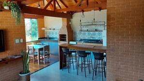 A spacious kitchen with a brick wall, wooden ceiling beams, and a built-in pizza oven. There are bar stools and a dining table with chairs. - Villa da Serra Ibitipoca (Lima Duarte)