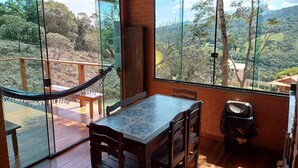 A room with a wooden table and chairs, a hammock, and large windows offering a view of a mountainous landscape. - Villa da Serra Ibitipoca (Lima Duarte)