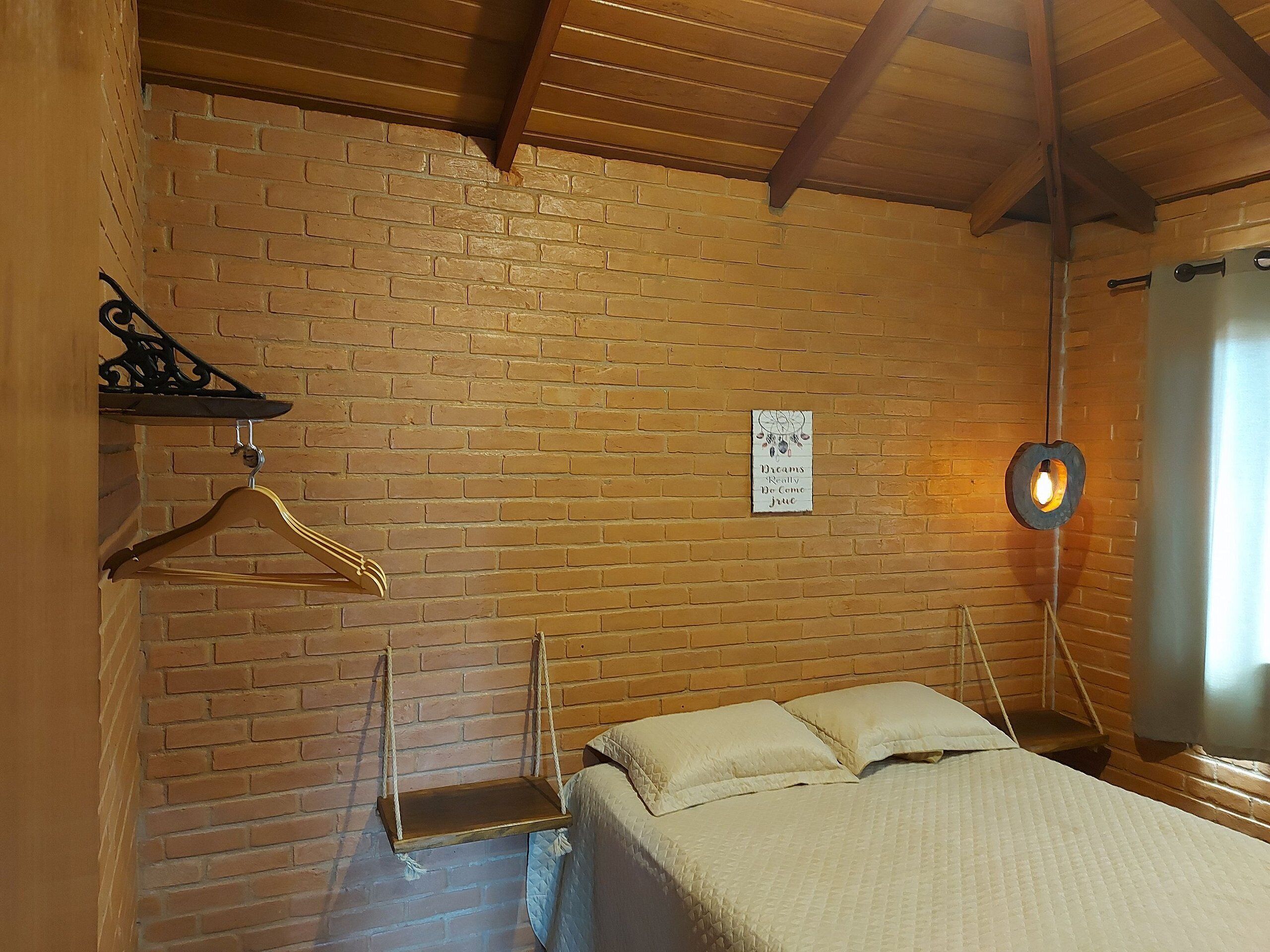 A room with a brick wall, a bed with pillows, a wooden hanger, a sign with text, and a hanging light fixture.