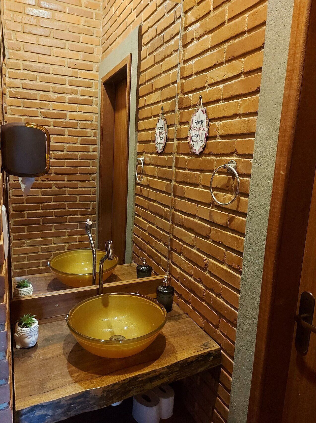 Un baño con pared de ladrillo, dos lavamanos con cuencos amarillos, un espejo, un perchero para toallas y un secador de manos.