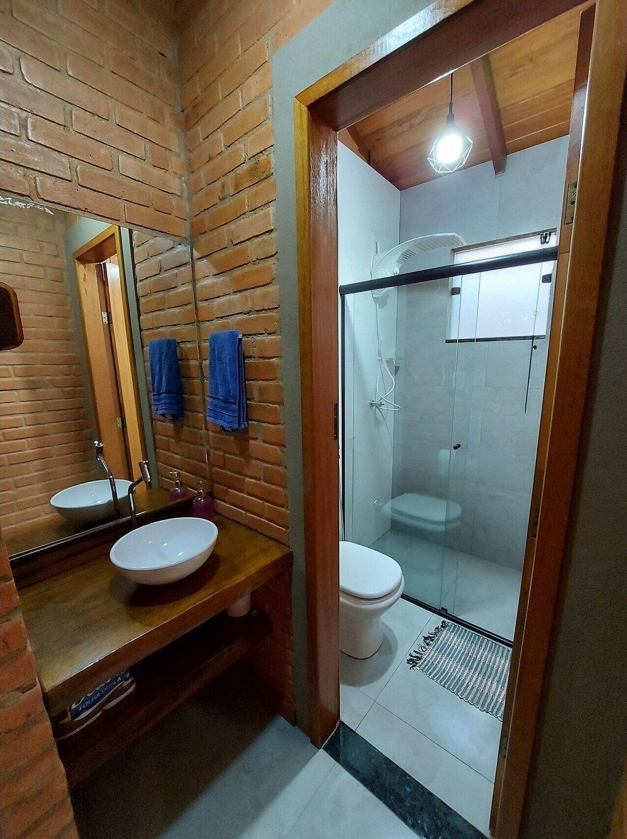 Un baño con pared de ladrillo, tocador de madera, dos lavamanos, un espejo y zona de ducha con mampara de vidrio.