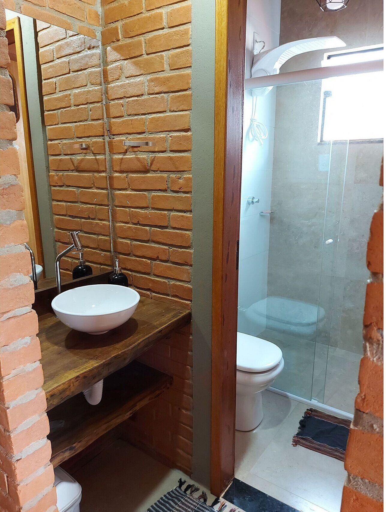 Een badkamer met een bakstenen muur, een houten wastafelmeubel, een witte wasbak, een toilet en een douchecabine met glazen wanden.