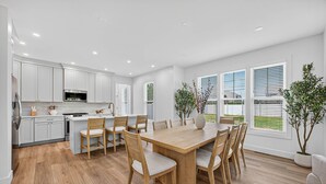 A modern kitchen with white cabinets, a wooden dining table, and chairs. There are potted plants and a view of the outdoors through large windows. - NEW BUILD - Walk to Notre Dame, Eddy Street Trader Joes only a half mile away (South Bend)