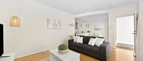 A modern living room with a grey sofa, white coffee table, and a small potted plant. A kitchen is visible in the background with a white countertop and various items on it.