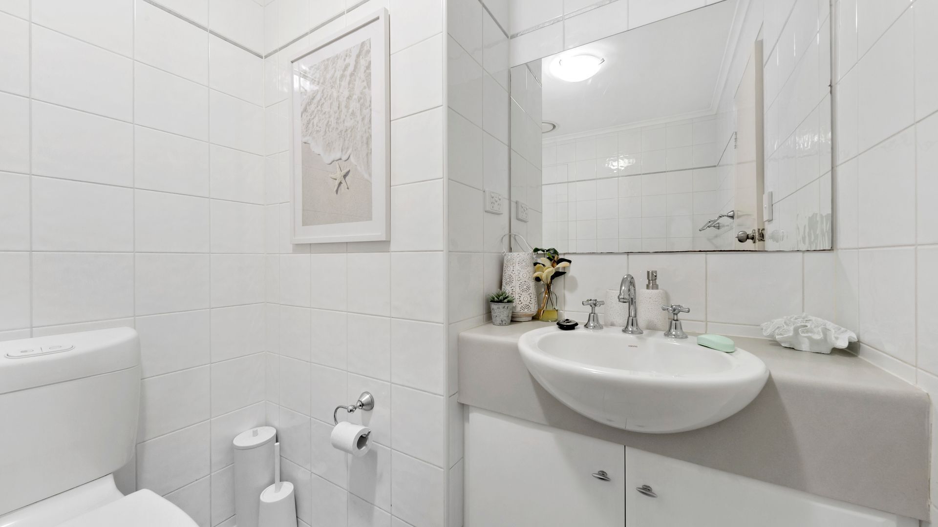 Ein modernes Badezimmer mit weißen Fliesen, einem großen Spiegel, einem Waschbecken mit mehreren Wasserhähnen und einer Toilette.