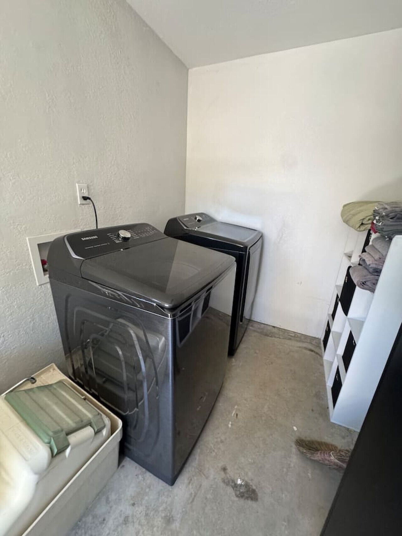 A compact laundry room with a front-loading washer and dryer, a small table, and a storage unit with folded clothes.