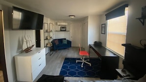 Habitación compacta con cama, escritorio con silla, televisor montado en la pared y ventana con persianas.