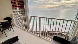 A balcony with wicker furniture, a small table with a plant, and a view of the ocean and buildings in the background.