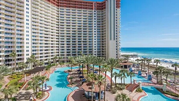 A high-rise hotel with a large swimming pool area, palm trees, and a beach view.