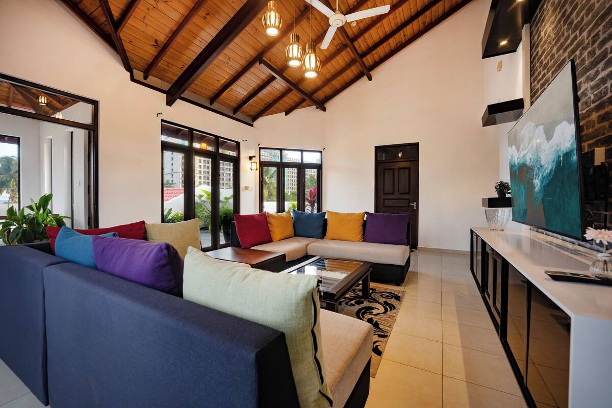 A modern living room with a sectional sofa, a glass coffee table, and a kitchen area with a large painting.