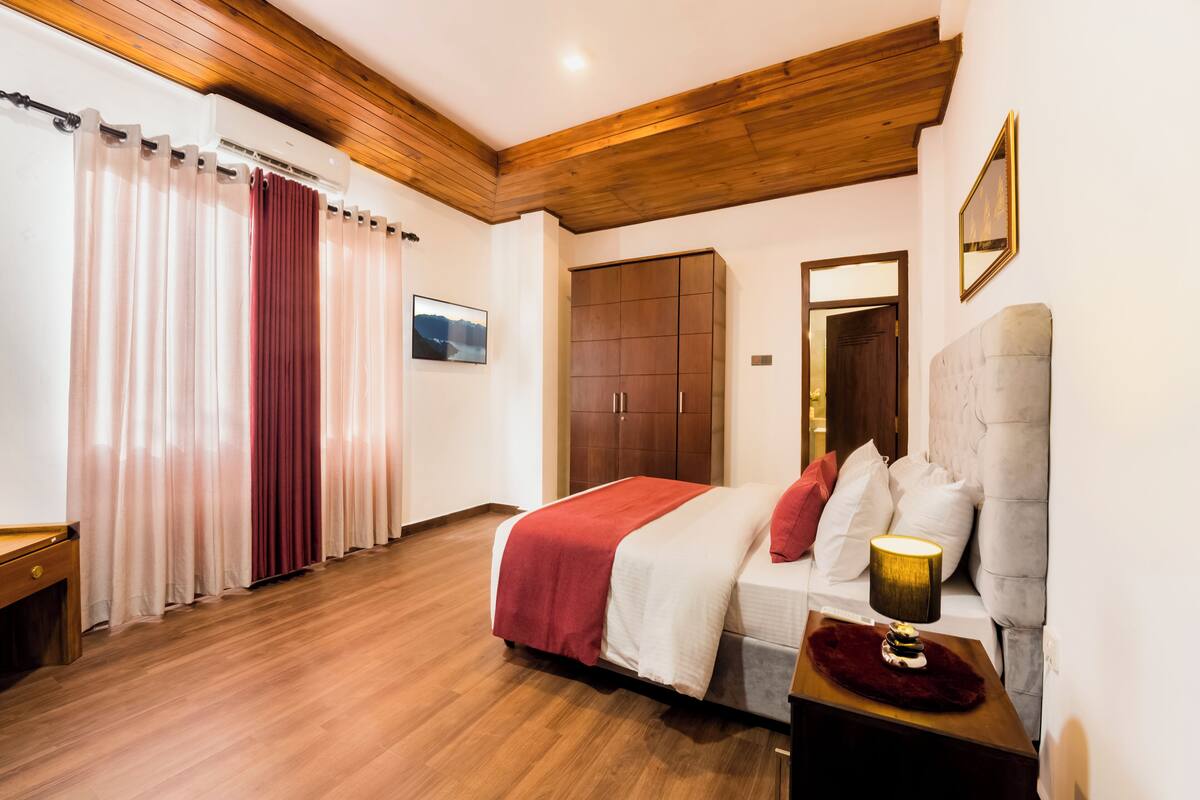 A bedroom with a wooden ceiling, a bed with a red and white bedspread, a nightstand with a lamp, and a wardrobe.