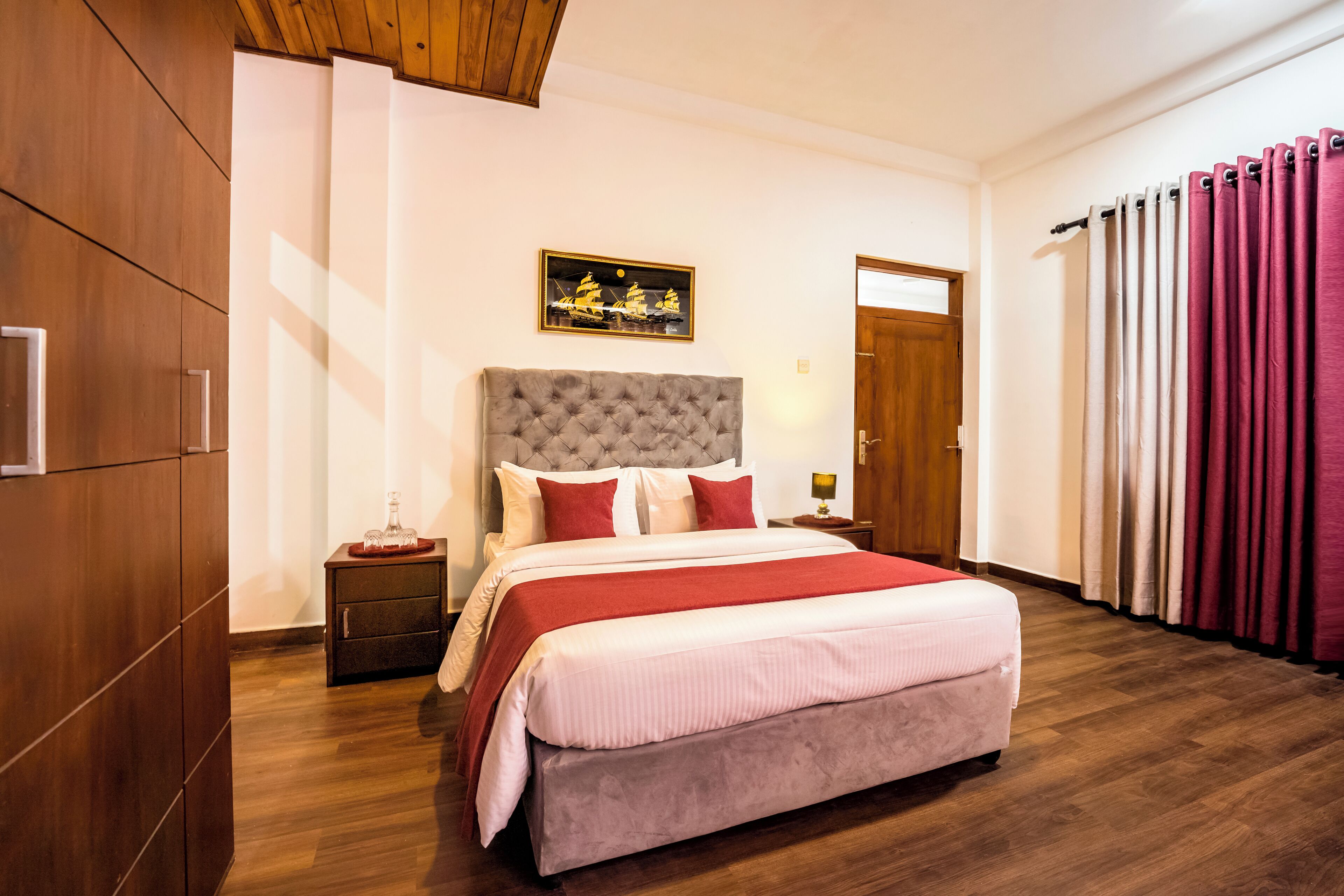 A hotel room with a large bed, wooden wardrobe, a nightstand with a lamp, and a door with curtains.