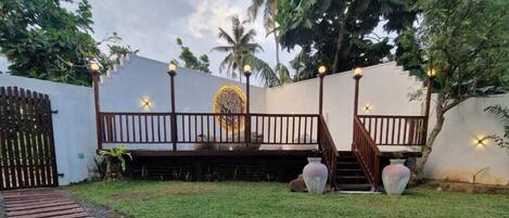 A wooden deck leading to a white wall with a decorative emblem, a staircase, and a paved area with pebbles.
