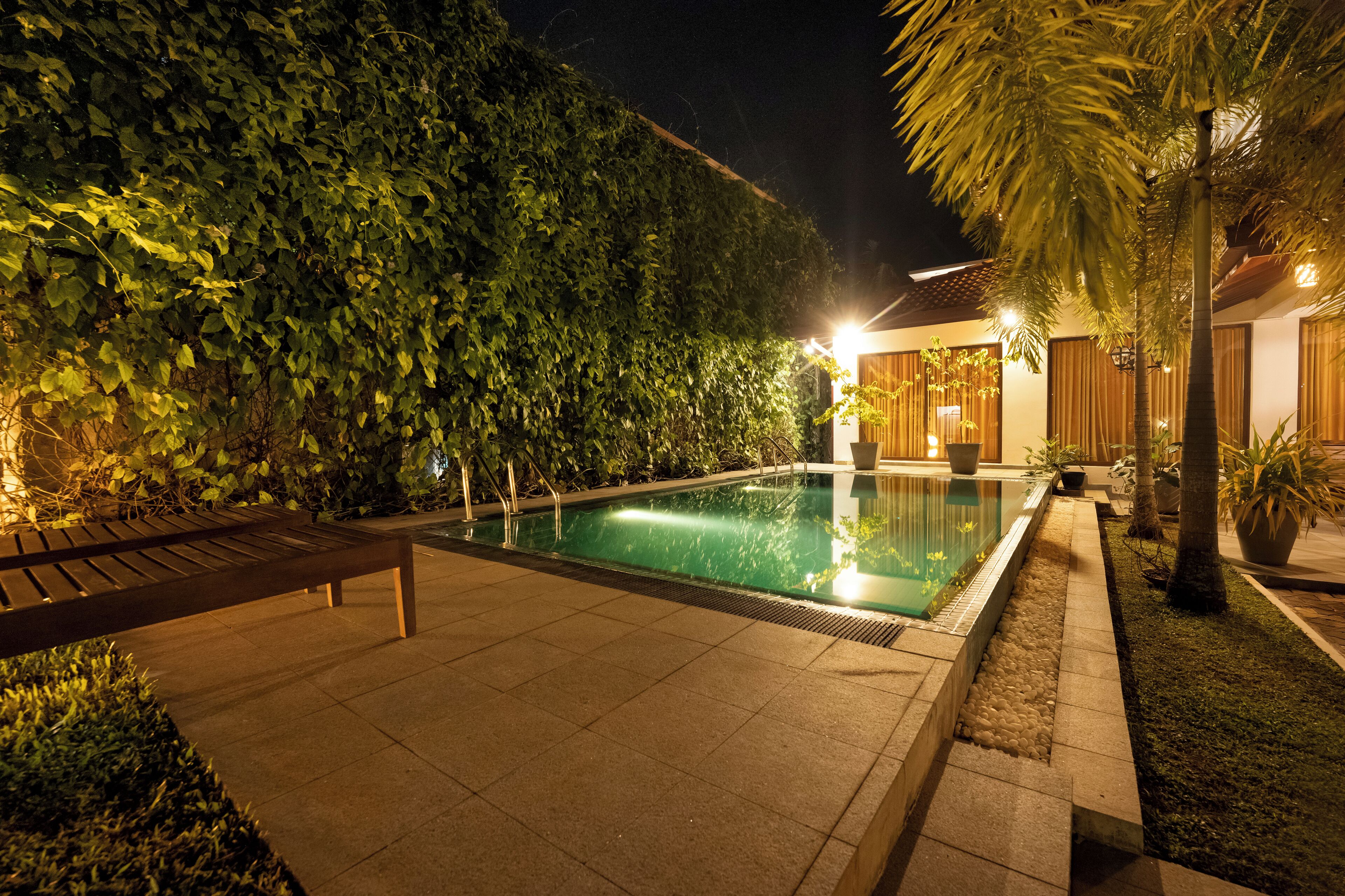 Un área de piscina nocturna con un banco, rodeada de exuberante vegetación y un edificio bien iluminado.
