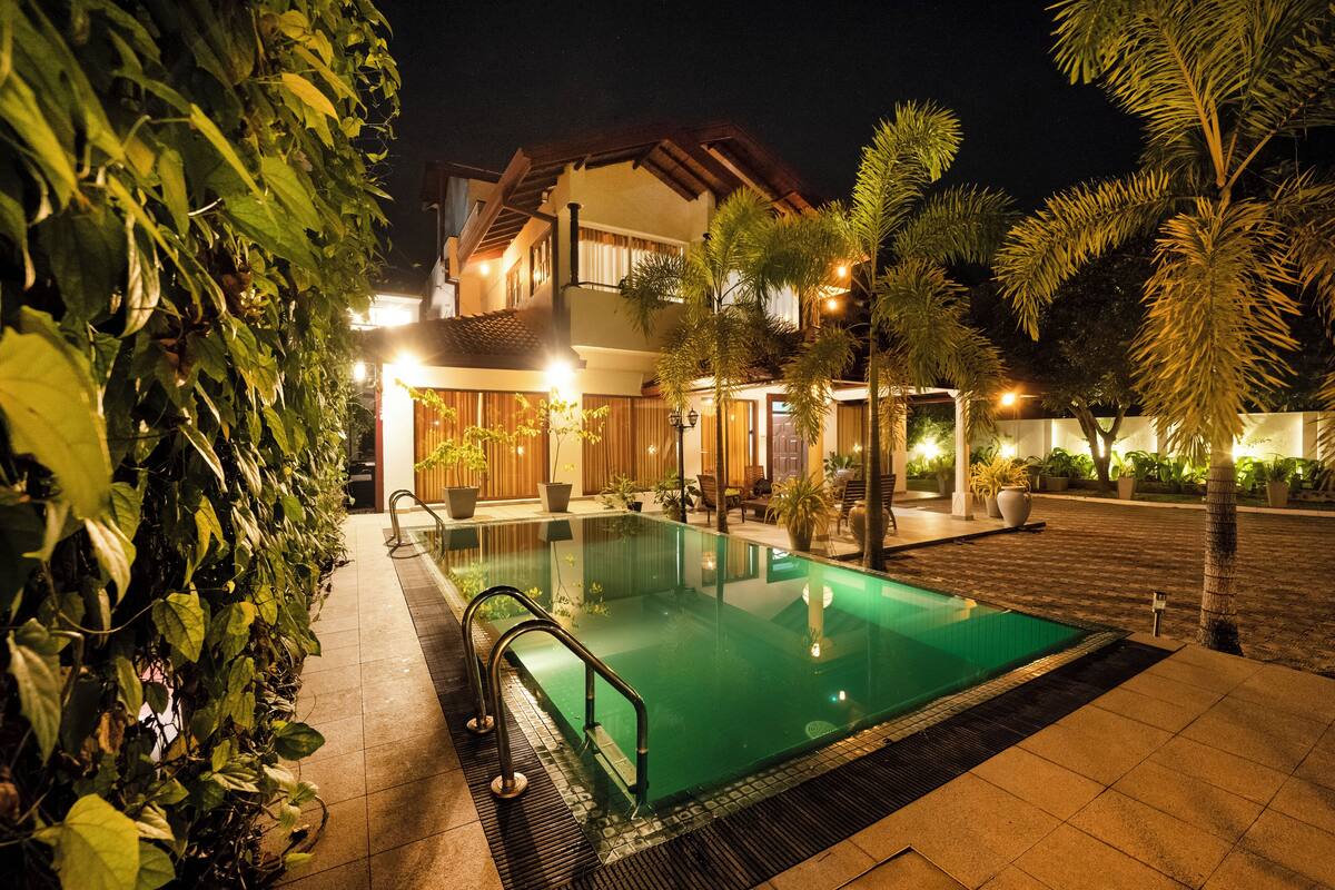 A swimming pool with a tiled deck, surrounded by potted plants and a building with a tiled roof.