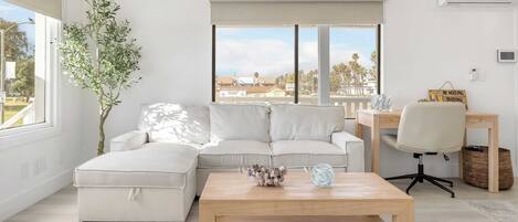 A modern living room with a sectional sofa, a wooden coffee table, a desk with a chair, and a large window with a view of palm trees and buildings.