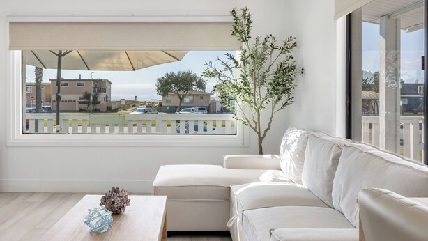 A modern living room with a sofa, a small table with decorative items, a large window with a view of buildings and a street, and a potted plant. - Walk to the Sand! Ocean Views, Balcony & Parking Sunset Surf (Huntington Beach)