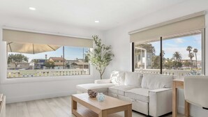 A modern living room with a white sofa, a wooden coffee table, and a large window with a view of palm trees and buildings. - Walk to the Sand! Ocean Views, Balcony & Parking Sunset Surf (Huntington Beach)