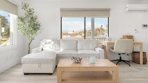 A modern living room with a sectional sofa, a wooden coffee table, a desk with a chair, and a large window with a view of buildings and palm trees. - Walk to the Sand! Ocean Views, Balcony & Parking Sunset Surf (Huntington Beach)