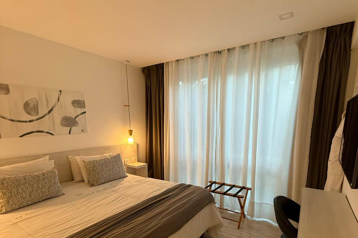 A hotel room with a large bed, a desk, and a chair. There is a window with curtains and a painting on the wall.