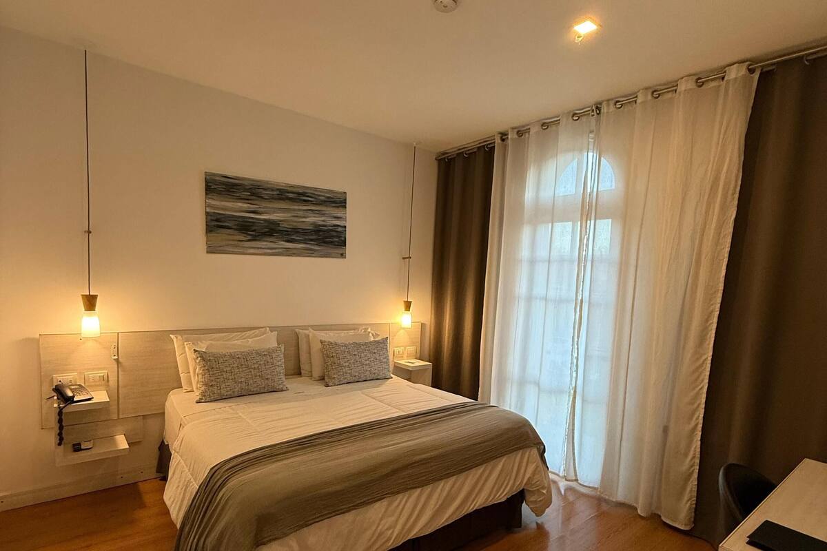 A bedroom with a large bed, bedside tables, a desk, and a window with curtains.
