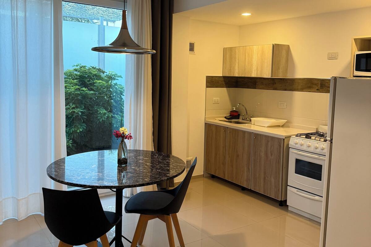 A modern kitchen with a dining area, featuring a round table, chairs, a hanging light fixture, and a view of greenery outside.
