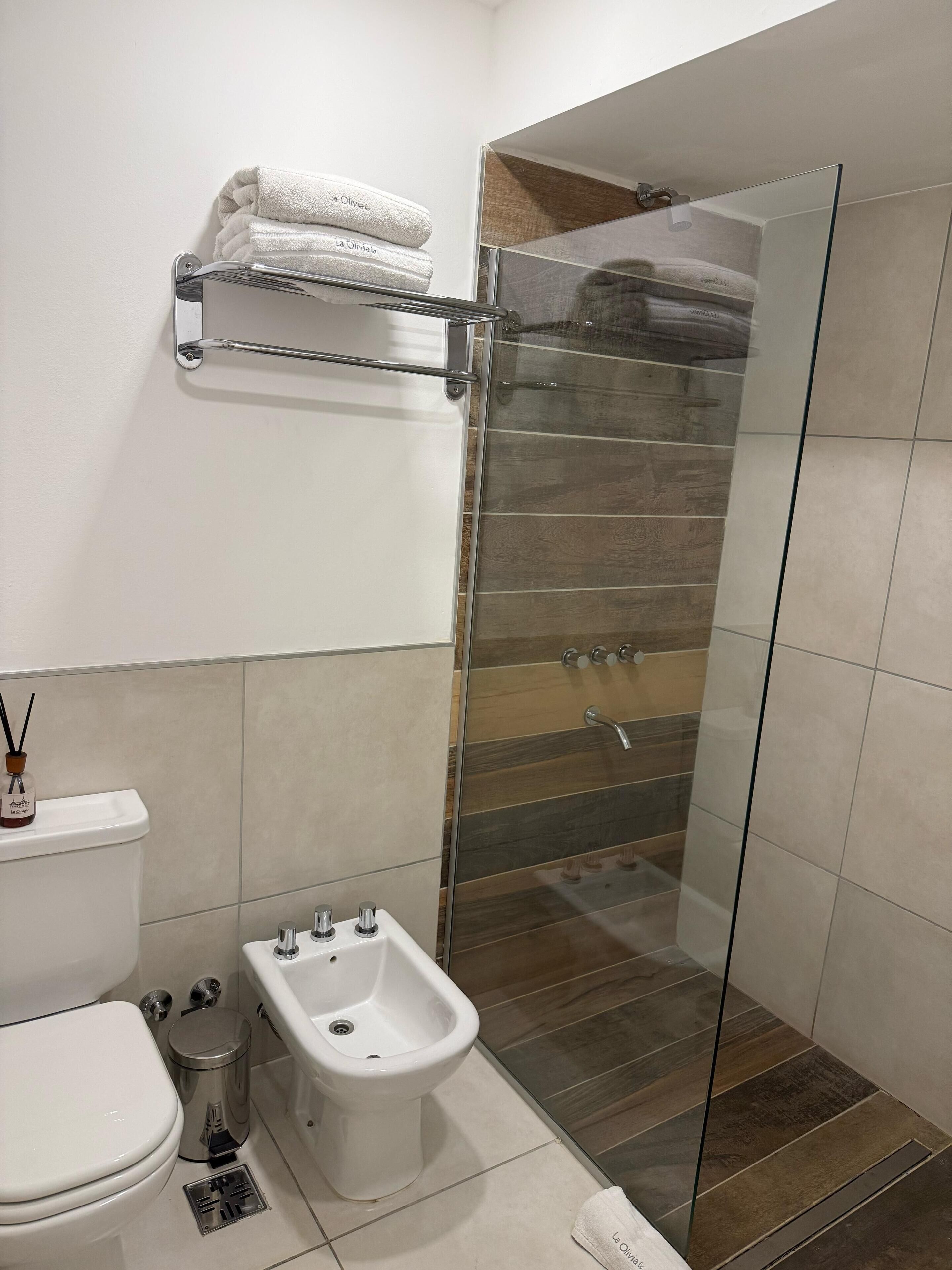 Un baño moderno con cabina de ducha de vidrio, un bidé, un inodoro y un estante con toallas dobladas.