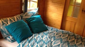 A wooden cabin room with a bed, two teal pillows, and a window with a view. - La roulotte du petit bois (Thénouville)