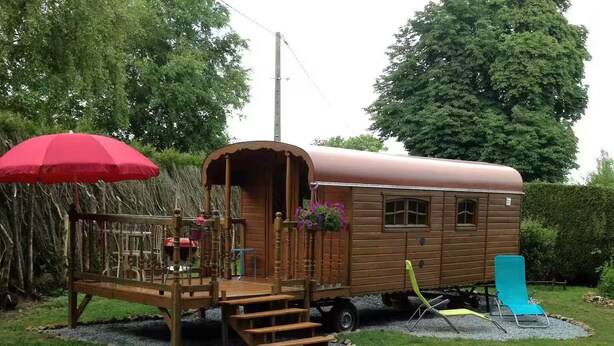 A wooden covered wagon with a porch, steps, and a small garden area with a red umbrella and a blue chair. - La roulotte du petit bois (Thénouville)