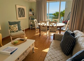 Een kamer met een bank, een eettafel met stoelen, een boekenkast en uitzicht op de oceaan.
