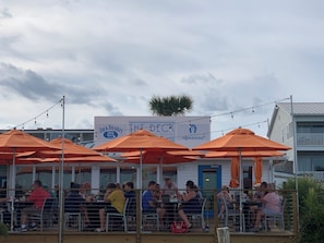 Un área de comedor al aire libre con sombrillas naranjas y mesas, un letrero que dice "The Deck" restaurante, y una vista de un edificio al fondo.