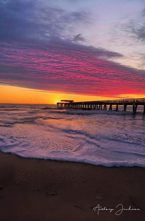 Una playa con un muelle que se adentra en el océano, olas mansas acariciando la orilla, y un atardecer con tonalidades rosadas y anaranjadas en el cielo.